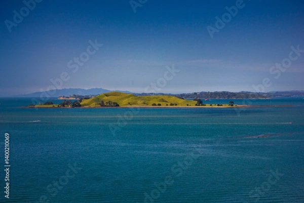 Fototapeta A stunning ocean view featuring a red ferry boat sailing near lush green islands in Auckland, New Zealand. The deep blue water contrasts beautifully with the vibrant landscape under a clear blue sky.