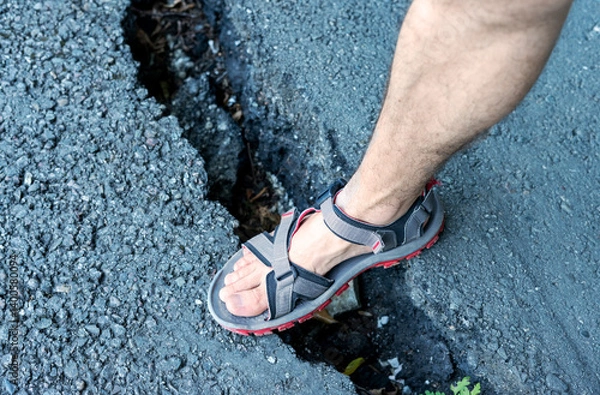 Fototapeta Foot in grey sandal steps into crack on an asphalt road, infrastructure damage, posing potential hazards for pedestrians Outdoor setting with visible gravel
