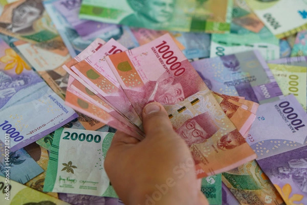 Fototapeta Hands holding several hundred thousand rupiah bills with a background of various types of Indonesian money. Suitable for background, finance, banking, cash, savings.