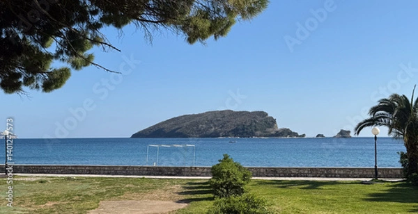 Fototapeta Scenic view of forested island surrounded by tranquil blue sea, framed by palm trees and coastal park.
