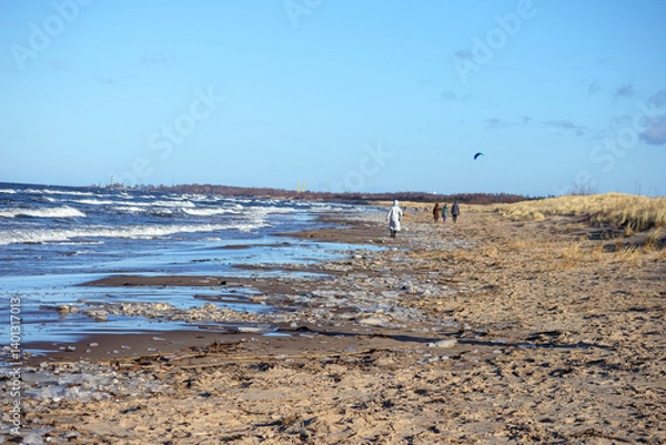 Fototapeta 
a sandy beach with a wavy sea, dry grass and three people walking further along the shore.