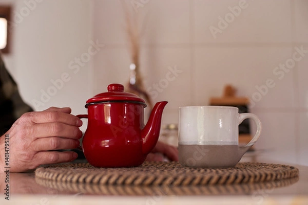 Fototapeta Elderly man gently holding a vintage red teapot, preparing a comforting morning tea ritual, with a ceramic mug placed nearby on a woven mat in a cozy kitchen setting