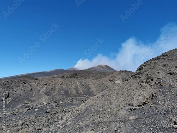 Fototapeta Rocky volcanic terrain with scattered lava stones and slopes surrounding a smoking crater on Mount Etna beneath a vivid blue sky in Sicily, Italy
