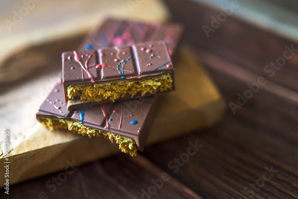 Fototapeta Close-up of artisanal Dubai chocolate bar with colorful topping, broken to reveal rich, golden filling on a rustic wooden board.