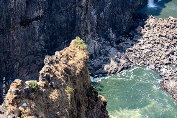 Fototapeta Rocky outpost in the victoria falls canyon with sun shining on it and the green rapids below 