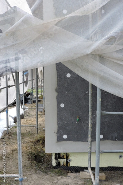 Fototapeta A wall of a house covered with EPS graphite polystyrene boards for thermal insulation, a scaffolding and building safety scaffolding net, some adhesive, anchors, an exterior wall mounted water faucet