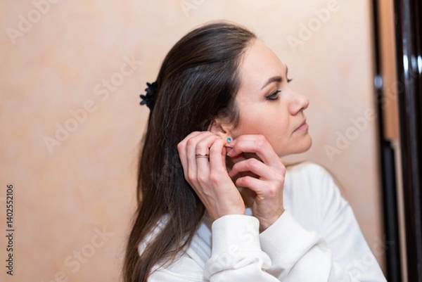 Fototapeta A young woman with long dark blond hair puts on an earring with a blue stone in her ear in front of a mirror..