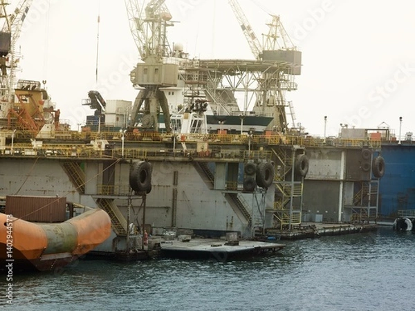 Fototapeta An offshore oil platform docked at a shipyard with cranes, support vessels, containers, and workers. The structure features yellow railings, massive tires, and maintenance crew on deck.