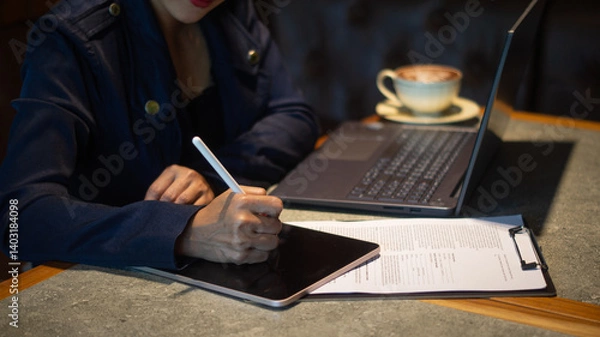 Obraz A businesswoman is intently writing something on a tablet placed on a checklist, using a white touchscreen pen, and working on a laptop in a cozy corner of a cafe.