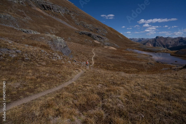 Fototapeta A man hiking through a vast rugged and desolated terrain of dry grass on a mountain slope, an alpine lake and blue sky, Ice Lake Trail, Weminuche Wilderness, San Juan National Forest, Colorado