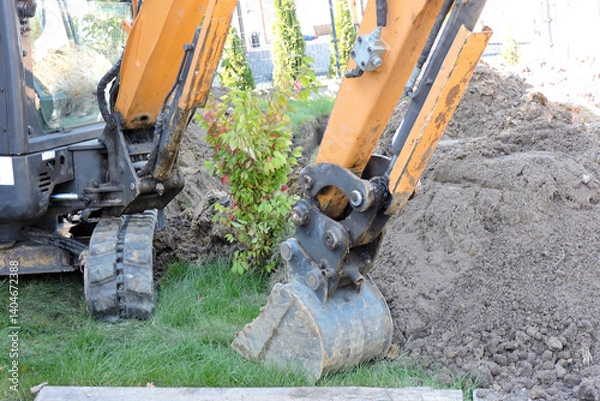 Fototapeta A narrow bucket, a dipper and hydraulic cylinders of a black and yellow mini hydraulic excavator standing in a courtyard next to heap of dirt