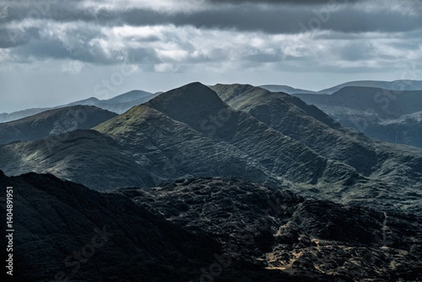 Obraz Dark, rolling hills under a cloudy sky. Shadows accentuate the rugged texture of the mountain slopes and valleys. Distant peaks are visible.