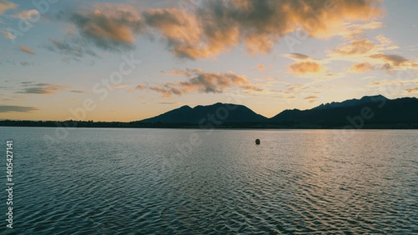 Fototapeta Scenic lake with mountains at sunset, reflecting a golden sky with scattered clouds.