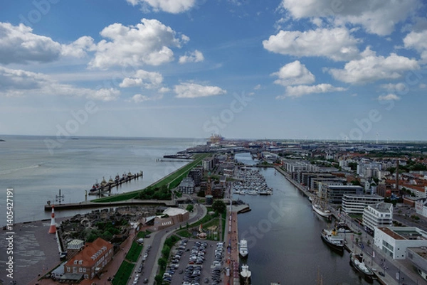 Fototapeta Coastal cityscape with modern buildings, docked ships, and a sandy beach under a partly cloudy sky.