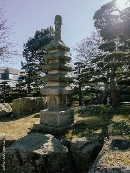Fototapeta A tall stone pagoda stands in a Japanese garden with manicured trees, rocks, and scattered purple flowers, under a bright sky with people in the background.