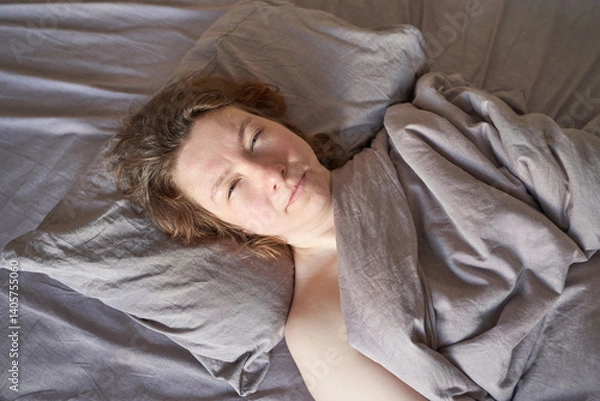 Fototapeta Caucasian female adult relaxing in bed with cozy grey bedding and natural light in home setting.