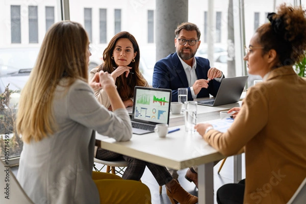 Fototapeta Businesswoman reviewing report on her laptop while discussing business growth with coworkers in meeting