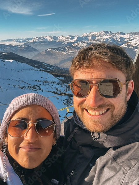 Fototapeta Smiling couple wearing sunglasses takes selfie with snowy mountain landscape in background