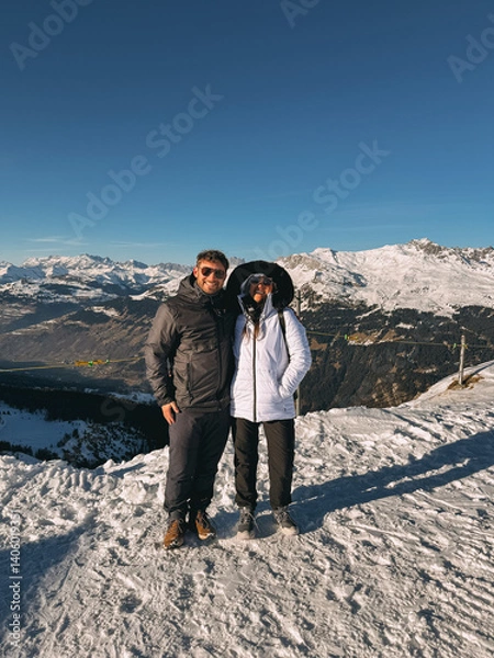 Fototapeta Two hikers stand on a snowy mountaintop, enjoying a breathtaking view of the alps