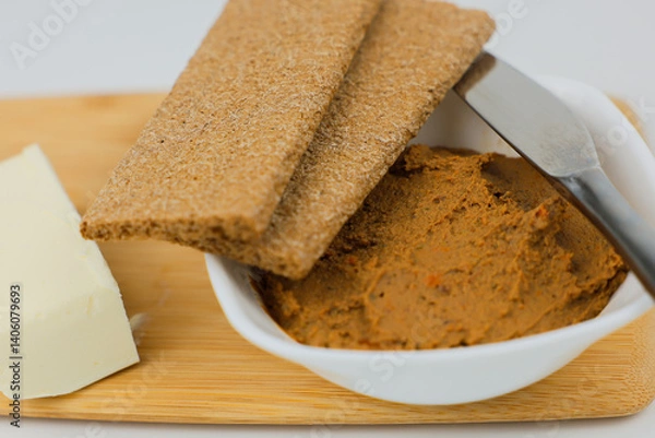 Obraz In close-up on a wooden board there is a white plate with liver pate, slices of bread and a knife lie on top and a piece of butter on a white background next to it. 