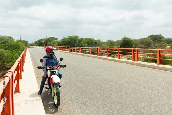 Fototapeta A man is riding a motorcycle on a road with a bridge in the background