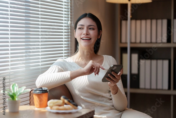 Obraz Cheerful young asian woman sitting by the window, enjoying breakfast with coffee and donuts while using her smartphone, embracing a cozy and relaxed morning at home