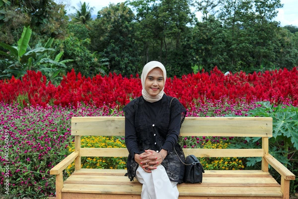Obraz A young woman wearing a hijab and casual outfit sits on a simple wooden park bench.