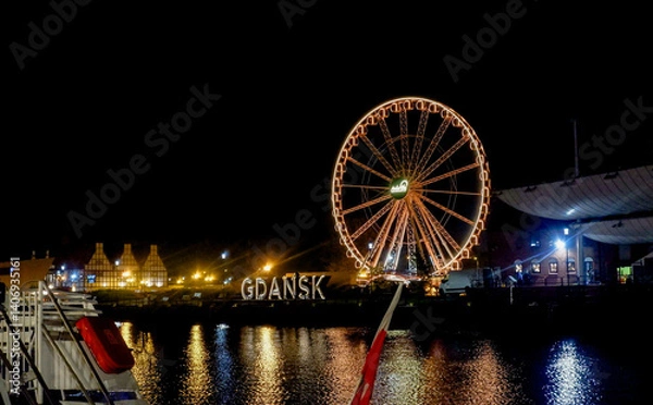 Fototapeta Night view of the illuminated Ferris wheel on Granary Island (Wyspa Spichrzów). The vibrant lights of the Ferris wheel reflect on the Motława River - "GDAŃSK" sign.
