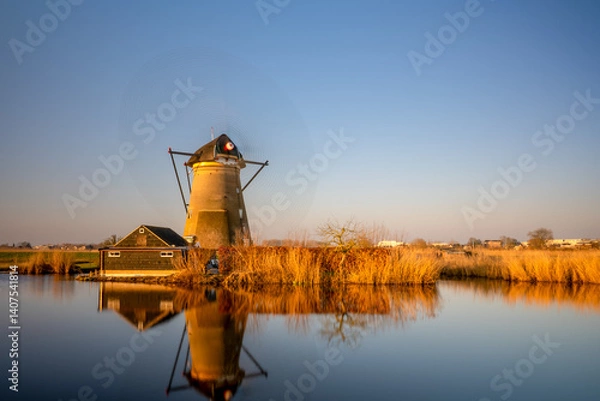 Fototapeta Traditional Dutch windmill reflecting on calm water during golden hour in the Netherlands.