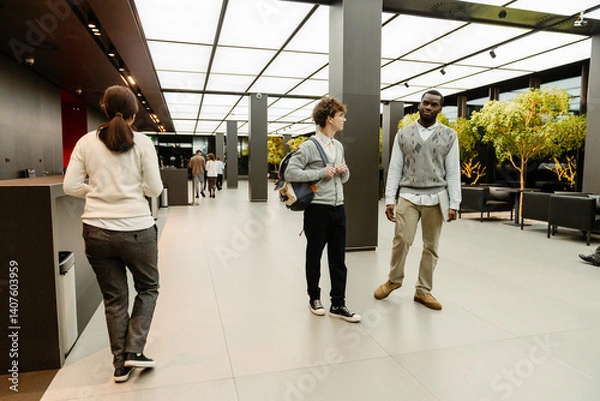 Fototapeta Two interns in their 20s, a White man in glasses and a cardigan with a backpack, and a Black man in a sweater vest with a laptop walk and talk in a modern office lobby with trees and other workers.