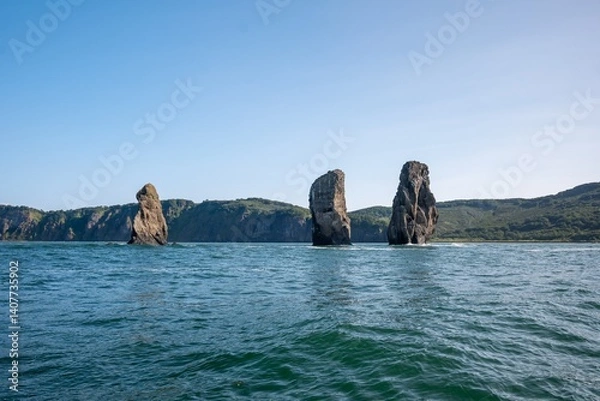 Fototapeta Three tall rock formations rising from the sea near a forested shoreline under a clear blue sky in a coastal landscape surrounded by calm waters and rugged terrain