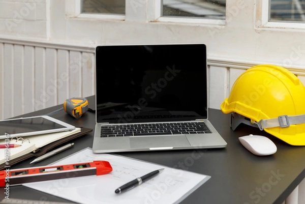 Obraz A laptop with a blank screen sits beside a yellow hard hat on a desk, suggesting a blend of technology and construction or engineering.