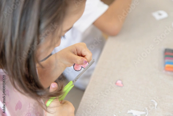 Fototapeta During a creative activity at home, a young child carefully uses safety scissors to cut out a heart shape, developing fine motor skills, autonomy, and concentration.