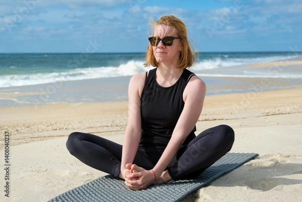 Obraz Woman practicing yoga on sandy beach, sitting in butterfly pose on mat, embracing mindfulness, relaxation, and wellness with ocean waves and blue sky background.