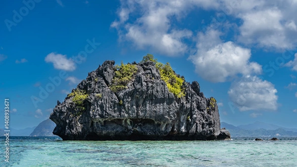 Fototapeta A rocky, lonely little island in a turquoise ocean. There is green vegetation on the steep, furrowed slopes. Blue sky, clouds. Philippines. Palawan. Bacuit bay