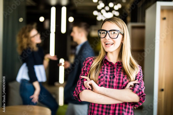 Fototapeta Portrait of a positive looking young woman standing with her arms crossed with coworkers talking in the background.