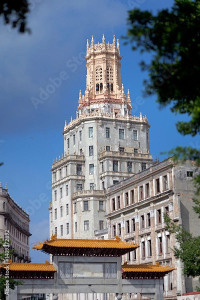 Obraz A historic ornate tower rises above the trees in Havana’s Chinatown (Barrio Chino), Cuba. The architecture blends neo-Gothic and Moorish elements with intricate stonework and slender arched windows.
