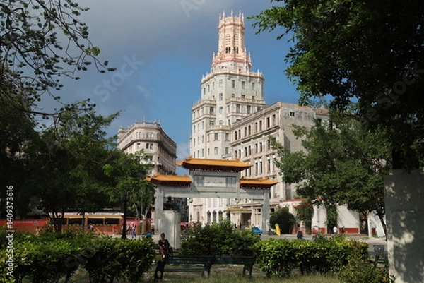 Obraz A historic ornate tower rises above the trees in Havana’s Chinatown (Barrio Chino), Cuba. The architecture blends neo-Gothic and Moorish elements with intricate stonework and slender arched windows.
