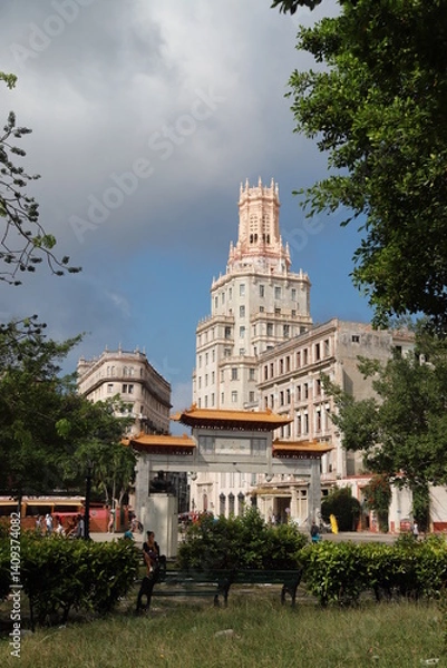Obraz A historic ornate tower rises above the trees in Havana’s Chinatown (Barrio Chino), Cuba. The architecture blends neo-Gothic and Moorish elements with intricate stonework and slender arched windows.
