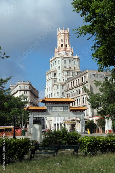 Obraz A historic ornate tower rises above the trees in Havana’s Chinatown (Barrio Chino), Cuba. The architecture blends neo-Gothic and Moorish elements with intricate stonework and slender arched windows.
