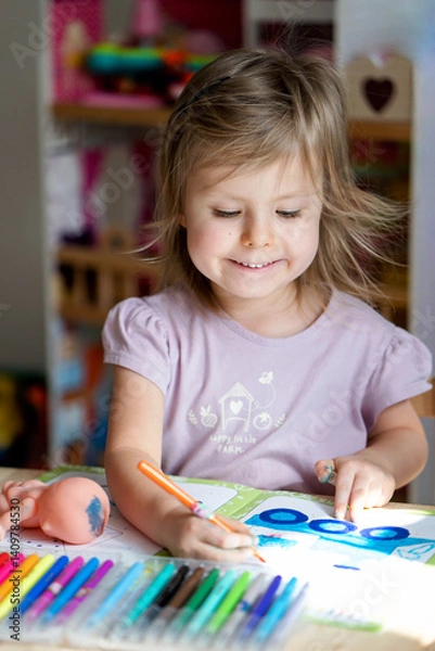 Fototapeta Happy child girl paints a drawing in an album and smiles at home or in kindergarten at the table. Creative little artist at work. A charming girl is drawing with felt-tip pens at the table.