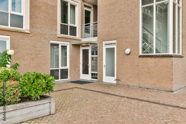 Fototapeta A contemporary building entrance showcasing large windows and a welcoming path lined with vibrant greenery, perfect for architectural appreciation.