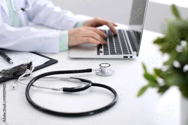 Fototapeta Close-up of stethoscope is lying on the table near female doctor typing on laptop computer