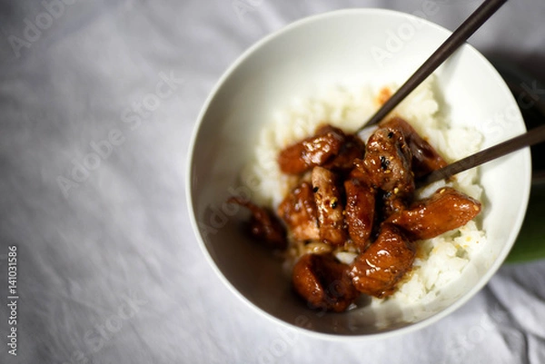 Fototapeta Plated chicken teriyaki over steamed white Jasmine rice in white ceramic bowl with wood chopstick on white tablecloth background
