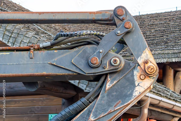 Fototapeta An excavator arm with visible hydraulic cylinders and structural elements against the background of a wooden building with a shingled roof. Traces of rust and wear are visible