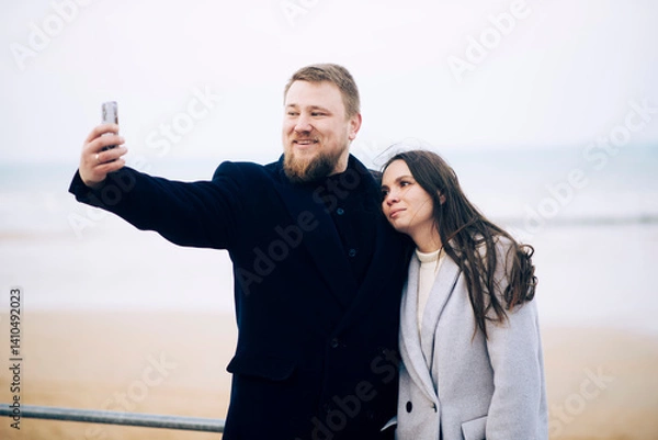 Obraz Young caucasian couple taking selfie by the beach on a cloudy day
