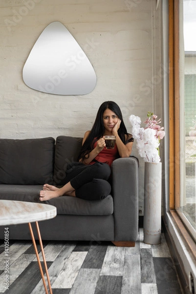 Obraz A woman sits on her sofa, legs crossed, enjoying coffee in her cozy living room, lost in thought, surrounded by a peaceful home atmosphere.


