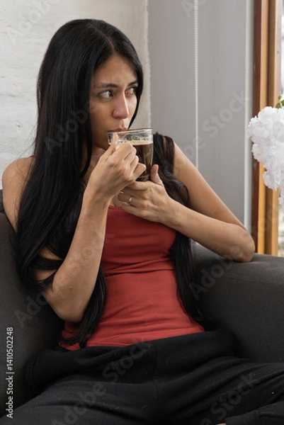 Obraz A young woman in a red blouse and black pants sits on a black sofa, holding a warm cup of coffee with both hands, relaxed and content.

