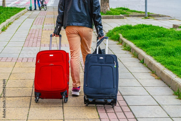 Obraz Traveler walking on sidewalk pulling red and black suitcases. Young man with two suitcases city street, person with luggage rear view. Travel mood, movement, modern lifestyle concept