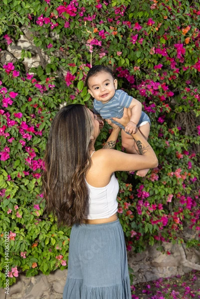Fototapeta Mother and son playing, mother holding a baby pointing to the sky, the baby is smiling and in the background there is a wall with plants, and both are wearing a cool outfit